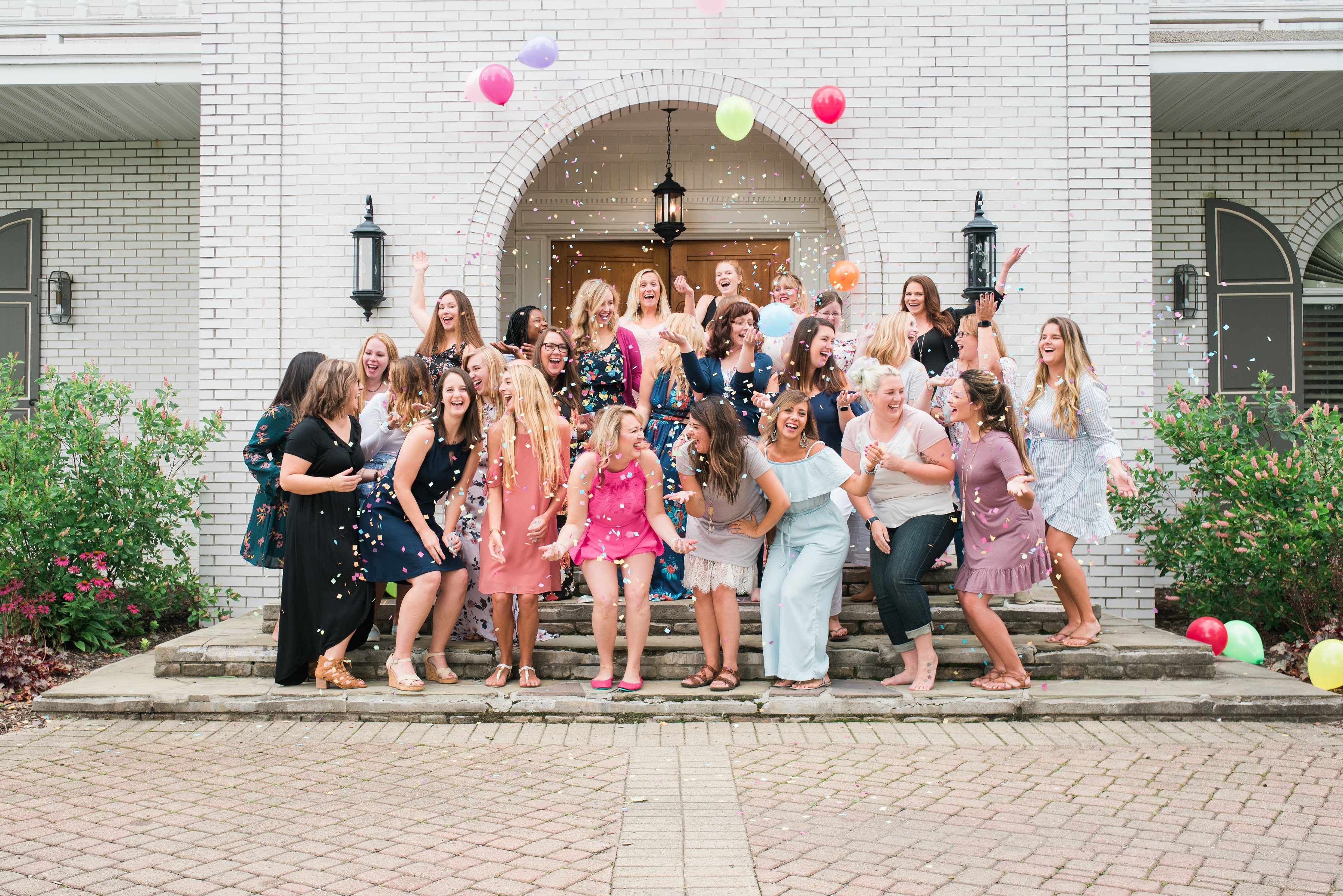 a large group of girls standing on the steps of a mansion throwing confetti and balloons