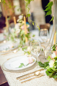 A tablescape setup at the George Peabody Library in Baltimore, MD shows off the gold dinner ware, gold tipped wine glasses, gold candles, and fine China surrounded by beautiful greenery and flowers 