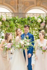 The wedding party stands laughing in front of a styled floral backdrop inside the George Peabody Library in Baltimore, MD