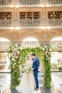 A bride and groom stand in front of a styled floral backdrop inside the George Peabody Library in Baltimore, MD sharing a newly wed kiss