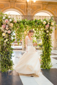 A bride twirls in her wedding dress in front of a styled floral backdrop inside the George Peabody Library in Baltimore, MD