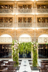 A styled floral backdrop sits at the back of a Ceremony inside the George Peabody Library in Baltimore, MD