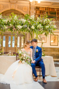 A bride and groom sit together in an intimate embrace in front of a styled table in the George Peabody Library in Baltimore, MD