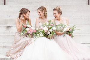 A bride and her bridesmaids sit on the steps of the George Peabody Library in Baltimore, MD laughing and having fun