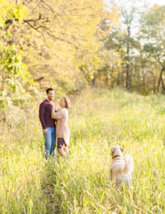 A September backyard anniversary session in Woodsboro MD. A couple stands together looking at each other lovingly in the tall grass under the glowy sun. Their dog joins