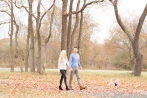 Cozy Autumn Engagement Session at Rockburn Branch Park in Elkridge, MD. A couple walks their dog in a park holding hands