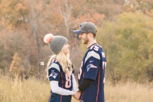 Cozy Autumn Engagement Session at Rockburn Branch Park in Elkridge, MD. A couple in Patriots Jerseys look at each other sweetly with orange fall leaves in the background