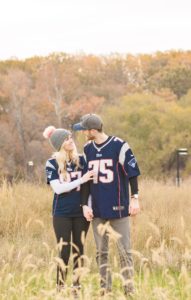 Cozy Autumn Engagement Session at Rockburn Branch Park in Elkridge, MD. A couple in Patriots Jerseys look at each other lovingly in a field with fall leaves in the background.