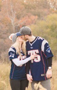 Cozy Autumn Engagement Session at Rockburn Branch Park in Elkridge, MD. A couple in Patriots Jerseys kisses in a field with fall leaves in the background.
