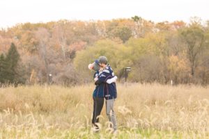 Cozy Autumn Engagement Session at Rockburn Branch Park in Elkridge, MD. A couple in Patriots Jerseys stands in a field with fall foliage in the background lovingly embracing.