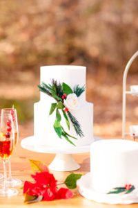 A Christmas themed wedding cake with white roses, pine needles and greenery created by the cake studio stands surrounded by Christmas desserrts, poinsettias, Christmas drinks and smaller holiday cakes at the Liriodendron Mansion in Bel Air Maryland for a winter wedding