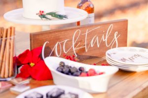 A cocktail sign made by the welcoming district stands on a winter dessert table surrounded by berries, plates, cinnamon sticks and drinks at the Liriodendron Mansion in Bel Air Maryland for a Christmas wedding