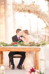 A young bride and groom sit at a sweetheart table draped in lace and evergreens smiling at each other lovingly beneath the wisteria of the Liriodendron Mansions back porch in Bel Air Maryland 
