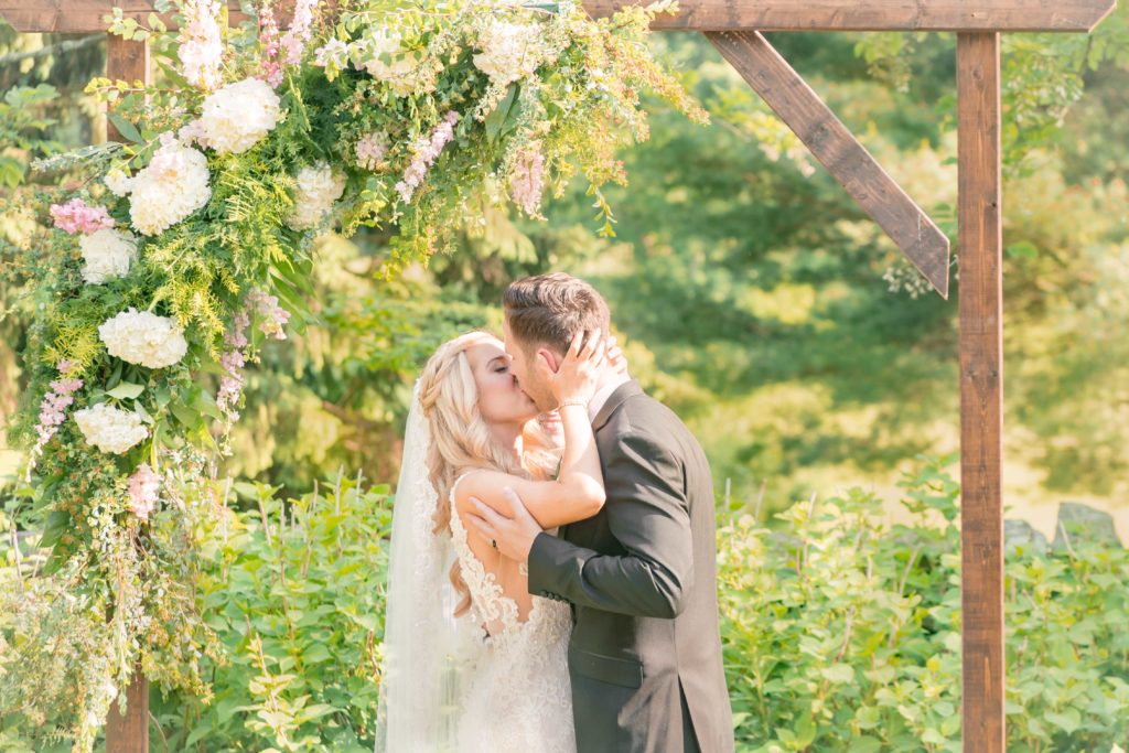 The Bride and groom stand underneath a flowering arch and share their first kiss in a summer garden wedding Airlie in Warrenton Virginia