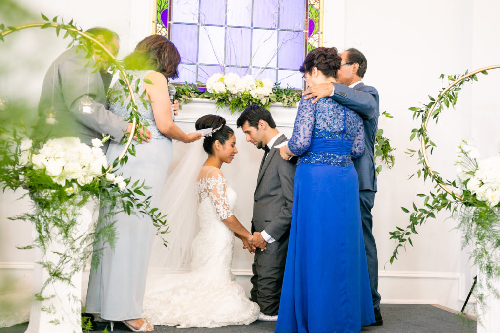 the Bride and groom kneel together in prayer with their parents standing over them in a latino wedding in the summer in frederick maryland wedding at the Seventh Day Adventist Church